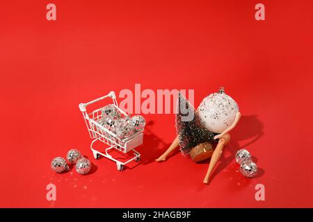 Holiday Shopping Thema weihnachtskugel mit Puppe Hände und Beine halten Tannenbaum in der Nähe Warenkorb mit Disco Kugeln auf rotem Hintergrund gefüllt. Kreati Stockfoto