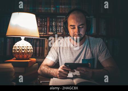 Lesen und Bildung. Ein Mann in der Bibliothek hört sich ein Hörbuch an, oder schaut sich ein Video am Telefon an, und es ist ein offenes Buch auf dem Tisch. Zoll Stockfoto