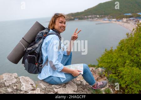 Lächelnde Touristenfrau, die auf einer felsigen Kante mit einer Karte in den Händen sitzt und für die Kamera posiert. Stockfoto