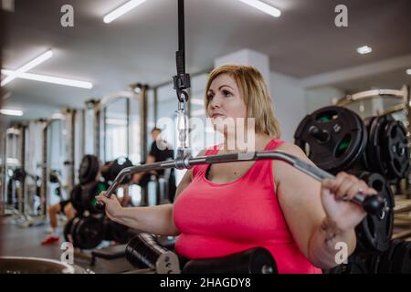 Mittlere Erwachsene plus große Frau, die im Fitnessstudio an einem Latenzausziehgerät trainieren kann Stockfoto