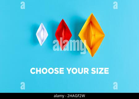 Drei Papierboote mit verschiedenen Größen und Farben auf blauem Hintergrund mit dem Text wählen Sie Ihre Größe. Stockfoto