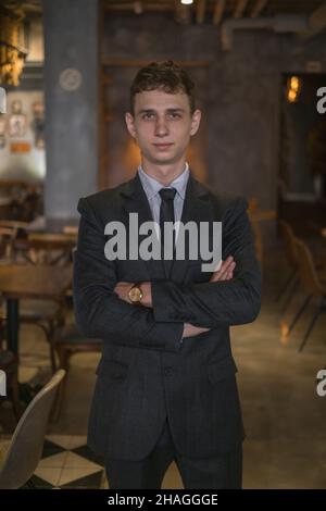 Porträt eines jungen kaukasischen Geschäftsmanns in formellem Anzug, der in der Bar mit gekreuzten Armen steht. Ein hübscher Mann, der im Café steht Stockfoto