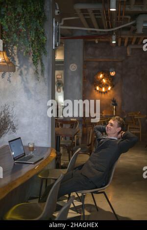 Schöner Mann, der in einem Café mit einem Laptop arbeitet. Online arbeitet, zufriedene Person, die sich im Café entspannt, erfolgreicher Abschluss des Arbeitstages Stockfoto