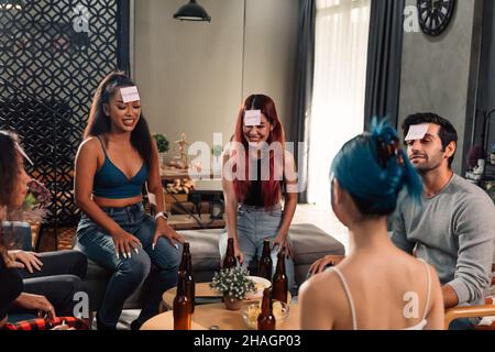 Gruppe von männlichen und weiblichen glücklichen Freunden, die Bier trinken und Spiel spielen, erraten, wer während des Spaßes zu Hause während einer Party und einem Wiedersehen Stockfoto