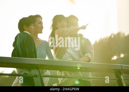 Asiatische Familie mit zwei Kindern, die auf einer Fußgängerbrücke stehen und die Aussicht im Stadtpark bei Sonnenuntergang genießen Stockfoto