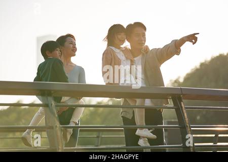 Asiatische Familie mit zwei Kindern, die in der Dämmerung auf einer Fußgängerbrücke stehen und die Aussicht im Stadtpark genießen Stockfoto