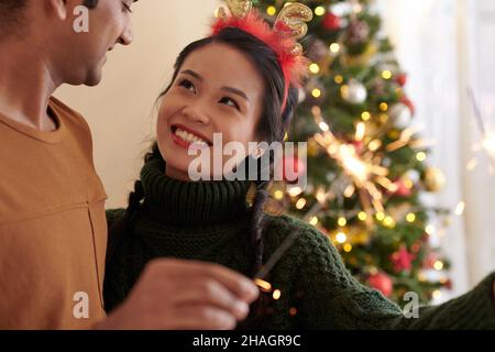 Hübsche lächelnde junge Frau, die in Liebe bengalisches Licht brennt und den Ehemann anschaut, wenn sie zu Hause Weihnachten feiern Stockfoto