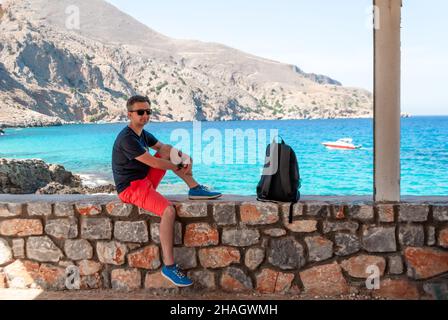 Ein Mann in rosa Aufnahmen, einem T-Shirt und einer Sonnenbrille sitzt auf einer Brüstung am Meer Stockfoto