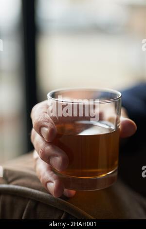 Nahaufnahme eines erfolgreichen Geschäftsmanns, der ein Glas alkoholischen Whiskys in der Hand hält, während er sich auf einem Stuhl entspannt Stockfoto