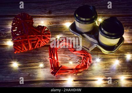 Auf einem hölzernen Hintergrund befinden sich rechts zwei Tassen Kaffee und links zwei große dekorative gewebte Herzen. Glühende Lichter sind um sich verstreut Stockfoto