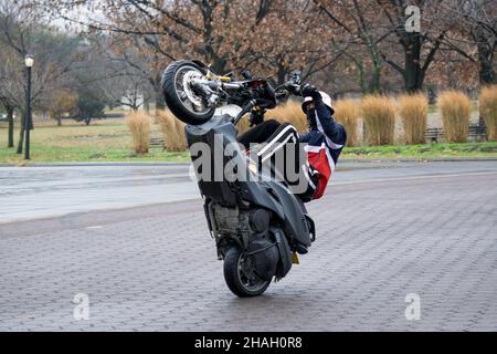 Ein Teenager-Biker mit Helm und Maske macht ein Wheelie auf seiner Yamaha TMax. In einem Park in Queens, New York City. Stockfoto