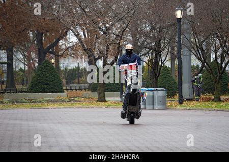 Ein Teenager-Biker mit Helm und Maske macht ein Wheelie auf seiner Yamaha TMax, während er auf dem Sitz steht. In einem Park in Queens, New York City. Stockfoto