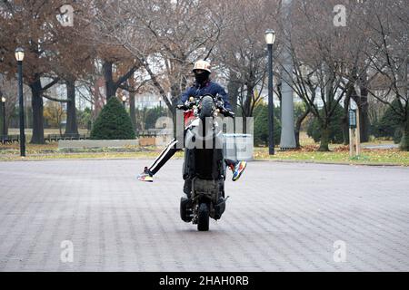 Ein Teenager-Biker mit Helm und Maske macht ein Wheelie auf seiner Yamaha TMax, während seine Beine vom Bike weg sind. In einem Park in Queens, New York City. Stockfoto