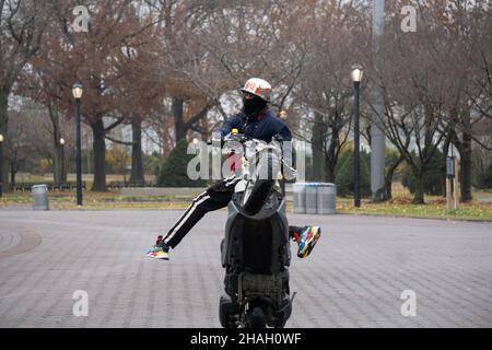 Ein Teenager-Biker mit Helm und Maske macht ein Wheelie auf seiner Yamaha TMax, während seine Beine vom Bike weg sind. In einem Park in Queens, New York City. Stockfoto