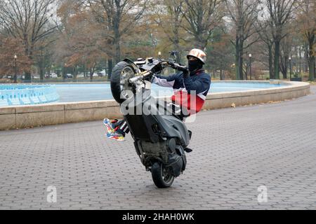 Ein Teenager-Biker mit Helm und Maske macht ein Wheelie auf seiner Yamaha TMax, während seine Beine vom Bike weg sind. In einem Park in Queens, New York City. Stockfoto