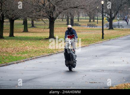 Ein Teenager-Biker mit Helm und Maske macht ein Wheelie auf seiner Yamaha TMax, während er auf dem Sitz steht. In einem Park in Queens, New York City. Stockfoto