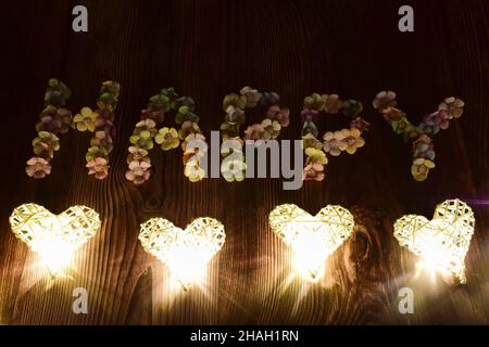 Auf einem hölzernen Hintergrund wird das Wort GLÜCKLICH in großen Buchstaben mit bunten Blumen geschrieben. Glühende Herzen sind in einer Linie von unten angeordnet Stockfoto