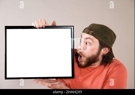Junger bärtiger Mann, der ein lustiges Gesicht macht und ein leeres Schild hält. Studio auf grauem Hintergrund aufgenommen. Stockfoto