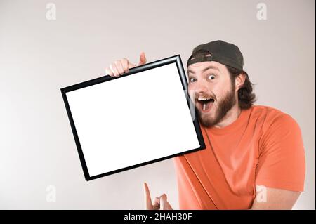 Junger bärtiger Mann, der ein lustiges Gesicht macht und ein leeres Schild hält. Studio auf grauem Hintergrund aufgenommen. Stockfoto