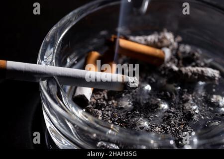 Nahaufnahme brennende Zigarette mit wunderschönem Rauchschwaden im Aschenbecher mit vielen Zigarettenkippen in vollem und schmutzigem Aschenbecher isoliert auf schwarzem Hintergrund CO Stockfoto