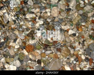 Meeressteine verschiedener Farben und Formen liegen unter dem klaren, klaren Wasser. Kreise von Tropfen breiten sich über das Wasser aus Stockfoto