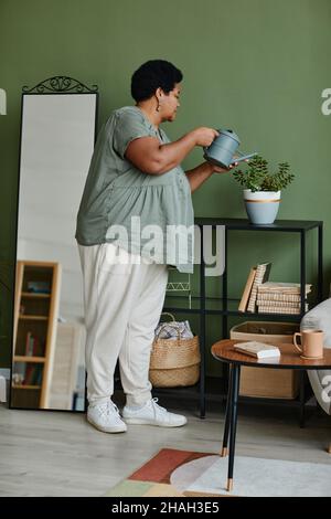 Vertikales Porträt einer schwarzen älteren Frau, die zu Hause Zimmerpflanzen wässert Stockfoto