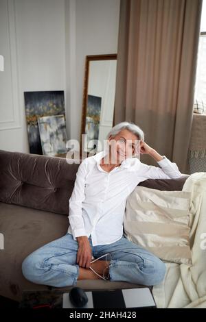 Ein hoher Winkel von charmanten Frauen mittleren Alters mit grauen Haaren und in Jeans und weißem Hemd, die mit gekreuzten Beinen auf der Couch zu Hause sitzen und vor der Kamera lächeln Stockfoto