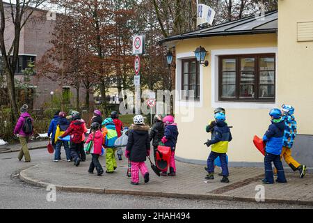 Eine Gruppe von Kindern, die Schneestürme aus Kunststoff tragen, überqueren in ihren bunten Kleidern vor und hinter einem Erzieher aus Sicherheitsgründen eine Straße. Stockfoto