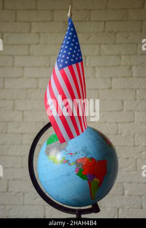 Die US-Flagge ist gegen einen verschwommenen Weltglobus mit obskuren Umrissen und einer Ziegelmauer aufgestellt. Unscharfer Hintergrund Stockfoto