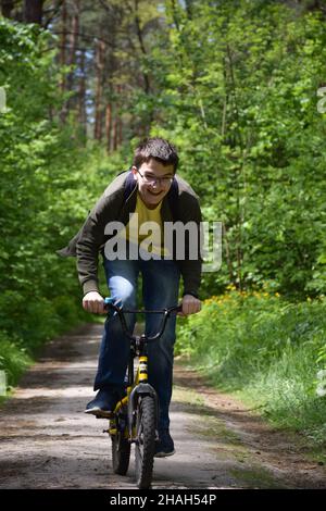 Ein etwa 14-jähriger Teenager, der eine Brille trägt, fährt schnell mit dem Kinderfahrrad einen Waldweg entlang und lacht. Vorderansicht Stockfoto