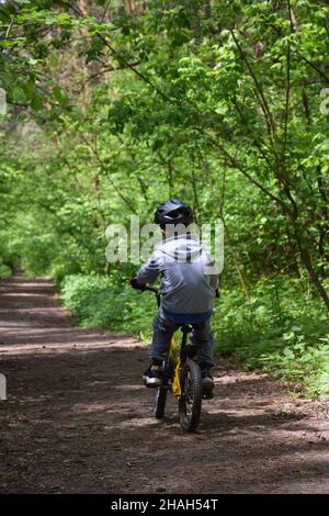 Ein etwa 8-jähriger Junge fährt mit einem gelben Fahrrad auf einem Waldweg in einem Sporthelm. Rückansicht Stockfoto