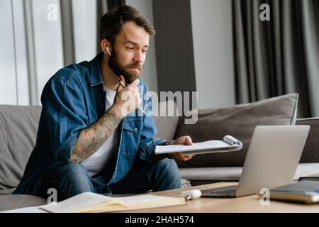 Bärtiger europäischer Mann, der mit einem Laptop arbeitet, während er zu Hause auf dem Sofa sitzt Stockfoto