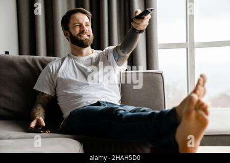 Bärtiger europäer, der lächelt und fernsieht, während er zu Hause auf dem Sofa sitzt Stockfoto