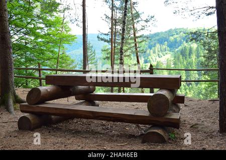 Auf dem Berg, im Wald, vor dem Hintergrund von Bäumen und einem anderen Berg, gibt es eine leere große Bank und einen handgefertigten Tisch aus Baumstämmen. Stockfoto