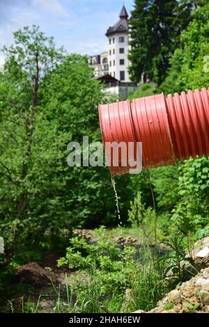 Aus einem roten Kunststoff-Abwasserrohr fließt ein dünner Wasserstrom vor einem verschwommenen Hintergrund aus Bäumen und einem Gebäude Stockfoto