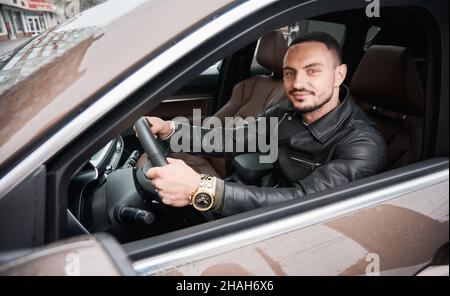 Porträt eines fröhlichen jungen Mannes, der hinter dem Steuer in seinem Auto sitzt. Kerl, der beide Hände auf das Lenkrad des geparkten Automobils legte und freundlich lächelte und die Kamera ansah. Stockfoto