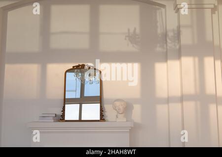 Spiegel auf künstlichen weißen Kamin in der Nähe von Statuette und Stapel von Büchern an der weißen Wand mit Schatten des Fensters und Kronleuchter darauf platziert Stockfoto