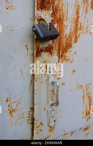 An einer Eisentür hängt ein Vorhängeschloss mit abblätternder Farbe und Rost. Ein einfacher eiserner Griff ist unter ihm sichtbar, nicht fokussiert Stockfoto