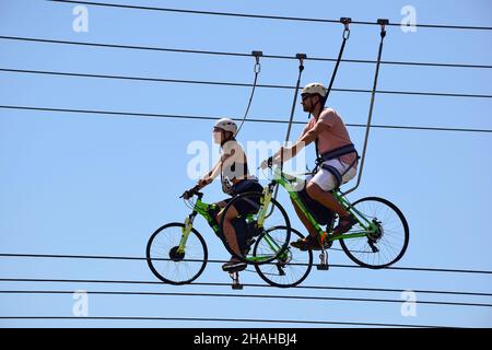 Bei der Attraktion fahren ein Mann und ein Mädchen auf Fahrrädern an Hängeseilen und lächeln. Von unten gegen den blauen Himmel fotografiert Stockfoto