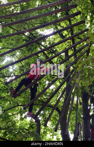 Ein Teenager in einem Sicherheitsgurt überquert eine Hängebrücke in einem Sommerpark Stockfoto