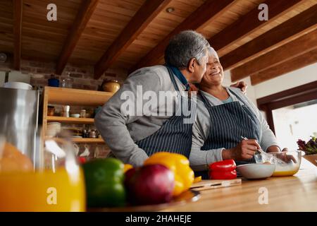Biracial ältere Paar glücklich Kochen in der Küche. Ehemann küsst Frau auf die Wange, gesunder Lebensstil im Ruhestand zu Hause. Stockfoto