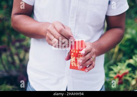 Die Hände eines Mannes öffnen einen roten Umschlag oder ein rotes Paket namens Ang Pao oder Ang Pow, auch Hongbao oder Hungbao auf Mandarin und Lai See auf Kantonesisch. Stockfoto