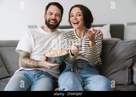 Glückliches junges Paar, das zu Hause auf einer Couch sitzt und fernsieht und Popcorn mit der Fernbedienung isst Stockfoto