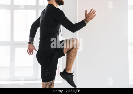 Junge Brünette bärtigen weißen Mann in Sportkleidung Ausübung springen drinnen Musik in Kopfhörer hören abgeschnitten Seitenansicht Stockfoto