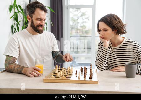 Glückliches junges weißes Paar, das Schach spielt und zu Hause am Tisch sitzt Stockfoto