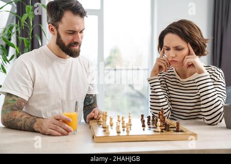 Glückliches junges weißes Paar, das Schach spielt und zu Hause am Tisch sitzt Stockfoto