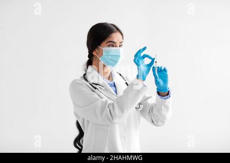 Seriöse junge indische Ärztin in Mantel, Schutzmaske und Handschuhen mit Spritze und Impfstoff auf weißem Hintergrund Stockfoto