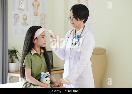 Lächelnder Arzt, der die Hand des präteen Mädchens hält, wenn er die Körpertemperatur mit einem berührungslosen Infrarot-Thermometer überprüft Stockfoto