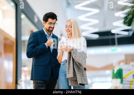 Glückliches, schönes junges Paar, das in der Halle des Einkaufszentrums mit dem Mobiltelefon steht. Bärtiger Mann in Brille und blonde Frau beim Kauf im Ladengeschäft. Stockfoto