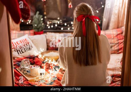 Gesichtslose Frau mit langen blondie Haaren und roten Bogen Haarclip Genuss zu Hause entspannen in gemütlichen bequemen Bett mit leuchtenden Lichtern Girlande Bokeh in der Nacht auf Stockfoto